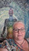 A woman with short gray hair and glasses wearing a colorful patterned top, standing in front of a large tapestry featuring a meditative figure and swirling mandala-like designs in blue, green, black, and cream.
