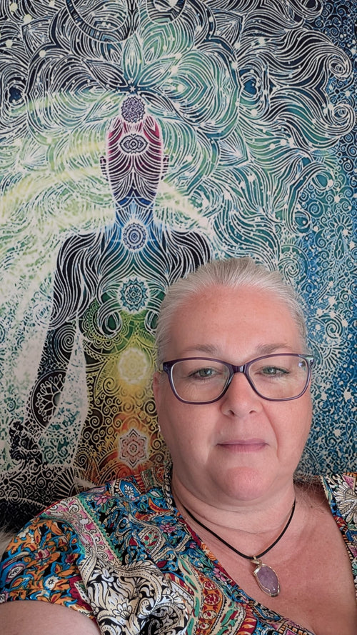 A woman with short gray hair and glasses wearing a colorful patterned top, standing in front of a large tapestry featuring a meditative figure and swirling mandala-like designs in blue, green, black, and cream.